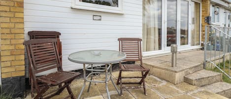 Eine Terrasse mit zwei Holzstühlen und einem Glastisch, eine Backsteinmauer und ein weißes Gebäude mit großen Fenstern und einer Tür.