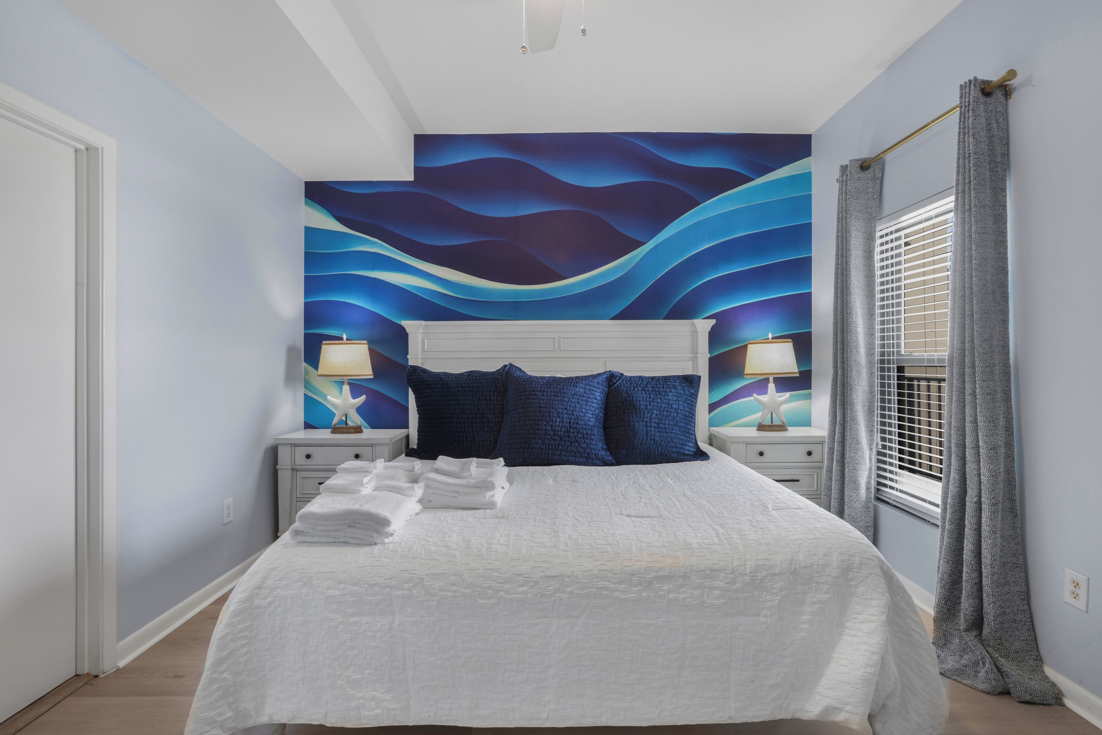 A bedroom with a bed, white headboard, two bedside tables with lamps, a window with curtains, and a wall mural.