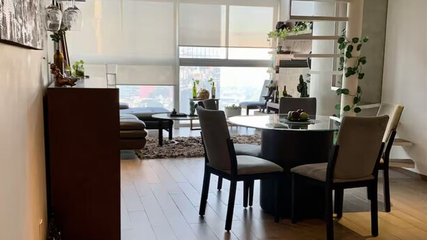 Un moderno comedor con una mesa redonda, dos sillas y vistas de la ciudad a través de grandes ventanales.