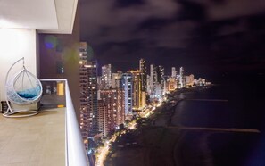 Un edificio alto con el horizonte urbano de noche, una silla colgante y una barandilla de balcón.