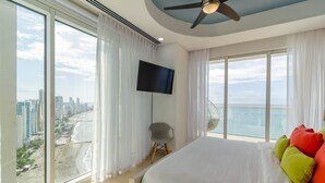 Quarto de hotel em um edifício alto, com uma cama grande, televisão e varanda com vista para a cidade e para a praia.