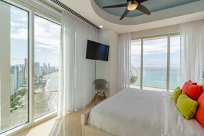 Ein Hotelzimmer in einem Hochhaus mit einem großen Bett, einem Fernseher und einem Balkon mit Blick auf Stadt und Strand.