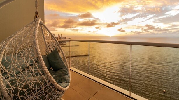 Varanda com cadeira suspensa, guarda-corpo de vidro e vista para a água e o pôr do sol.