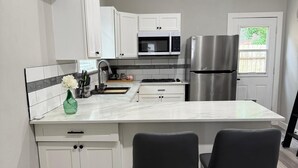 A modern kitchen with white cabinets, a stainless steel refrigerator, a microwave, and a gas stove. There are two black bar stools and a small green vase on the counter. - Private Guest House Free Parking 20min to IAH Airport (Houston)