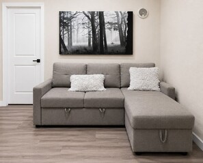 A grey sectional sofa with two white patterned cushions, a white door, and a forest-themed artwork on the wall.