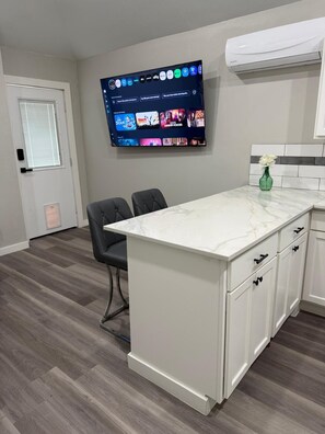 A modern kitchen with a marble countertop, white cabinetry, and a flat-screen TV mounted on the wall displaying various channels.