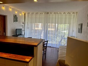 A room with a wooden table, a TV on a stand, a bed, a chair, and a window with sheer curtains. - El Tiburon N°17 - À 250 m de la plage (Argeles-sur-Mer)