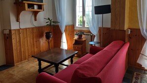 A living room with a wooden floor, a red sofa, a small table, a TV mounted on a wooden shelf, and two potted plants.