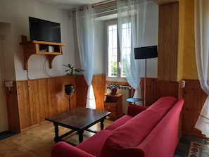A living room with a wooden floor, a red sofa, a small table, a TV mounted on a wooden shelf, and two potted plants. - Former San Giorgio Parish House between beautiful Lake Como and Lake Lugano (Pellio Intelvi)