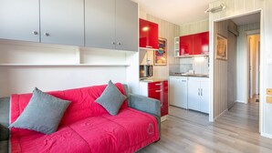 A modern kitchen with red cabinets, a grey sofa with grey cushions, and a wooden floor. - Horizon Montagne - À 150 m des Pistes de ski (Villarembert)