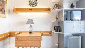 A compact room with a wooden table, a lamp, a clock, a radiator, and a television on a shelf. - Horizon Montagne - À 150 m des Pistes de ski (Villarembert)