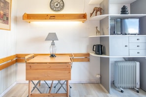 Habitación pequeña con una mesa de madera, una lámpara, un reloj, un radiador y un televisor sobre una estantería.