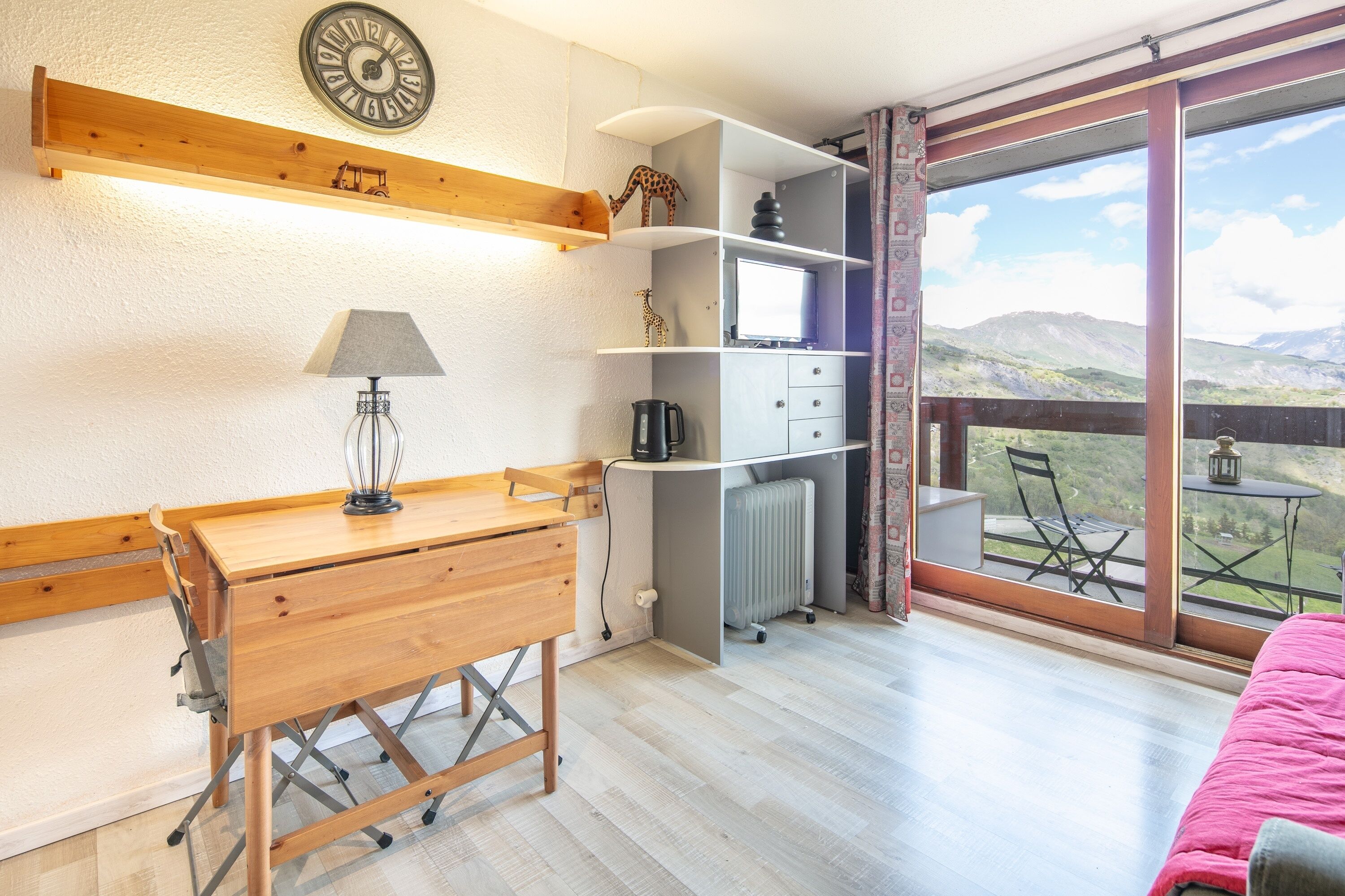 A room with a wooden desk, a lamp, a radiator, a television, and a large window with a view of hills.