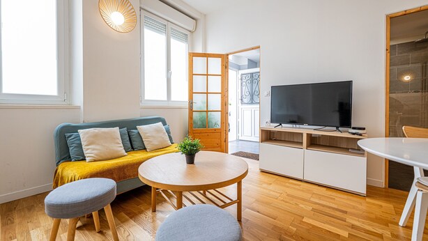 Ein modernes Wohnzimmer mit einer Couch, einem runden Holztisch, zwei Poufs, einem Fernseher und einer Küchenzeile mit Schränken.