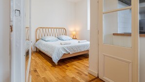 Ein Schlafzimmer mit einem Holzbett, einem Nachttisch, einer Lampe und Blick auf eine Küche durch eine Glastür.