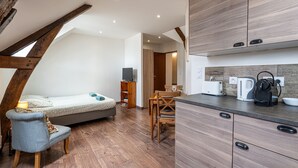 Een kleine keuken met houten kastjes, een eethoek met tafel en stoelen, een bed met hoofdbord en een ingebouwde televisie.