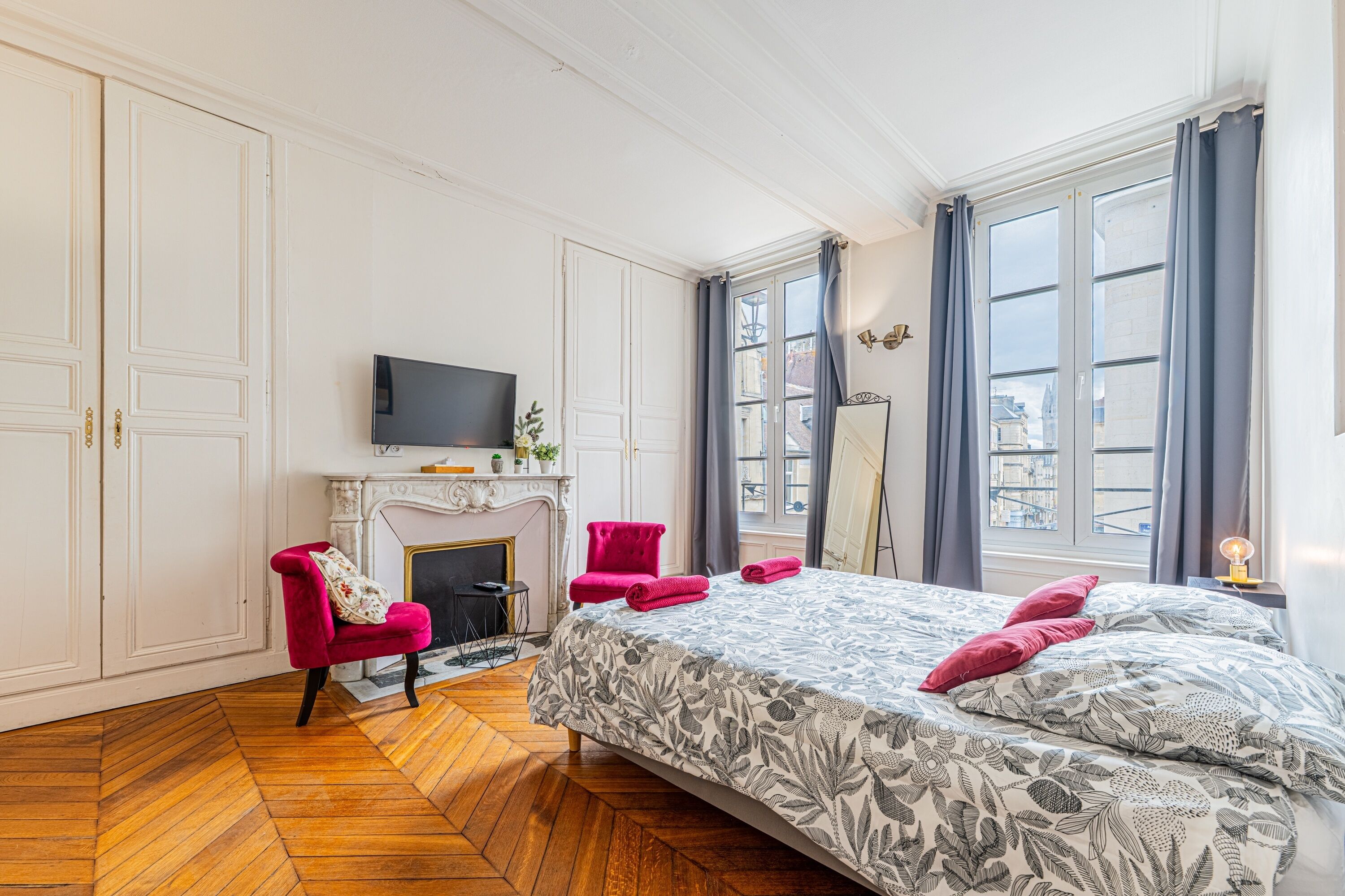 A hotel room with a large bed, a television, a fireplace, a red chair, and a view of the city through the windows.
