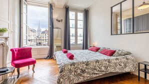 A room with a large bed, a red chair, a small table, and a view of buildings through a window.