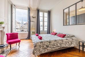 A room with a large bed, a red chair, a small table, and a view of buildings through a window.