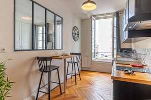 A kitchen with a wooden table, two chairs, a clock, a window, and a view of the outside.