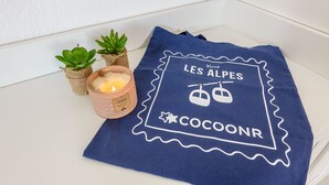 A blue towel with 'LES ALPES COCOONR' text and a mountain icon, placed on a white surface next to small potted plants and a lit candle. - Le Bel Air du Lac - Proche centre-ville d'Annecy (Annecy)