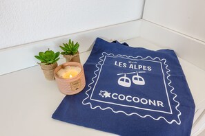 A blue towel with 'LES ALPES COCOONR' text and a mountain icon, placed on a white surface next to small potted plants and a lit candle. - Le Bel Air du Lac - Proche centre-ville d'Annecy (Annecy)