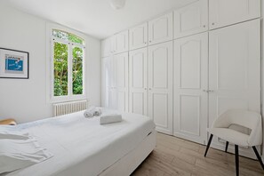 Ein Schlafzimmer mit Bett, Sessel, Fenster mit Grünblick und weißen Schränken.