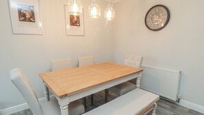 Ein Esszimmer mit Holztisch, weiß gepolsterten Stühlen, einer Bank, Pendelleuchten, einem gerahmten Bild und einer Wanduhr.