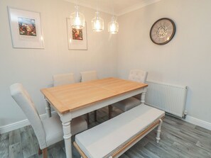 Ein Esszimmer mit Holztisch, weiß gepolsterten Stühlen, einer Bank, Pendelleuchten, einem gerahmten Bild und einer Wanduhr.