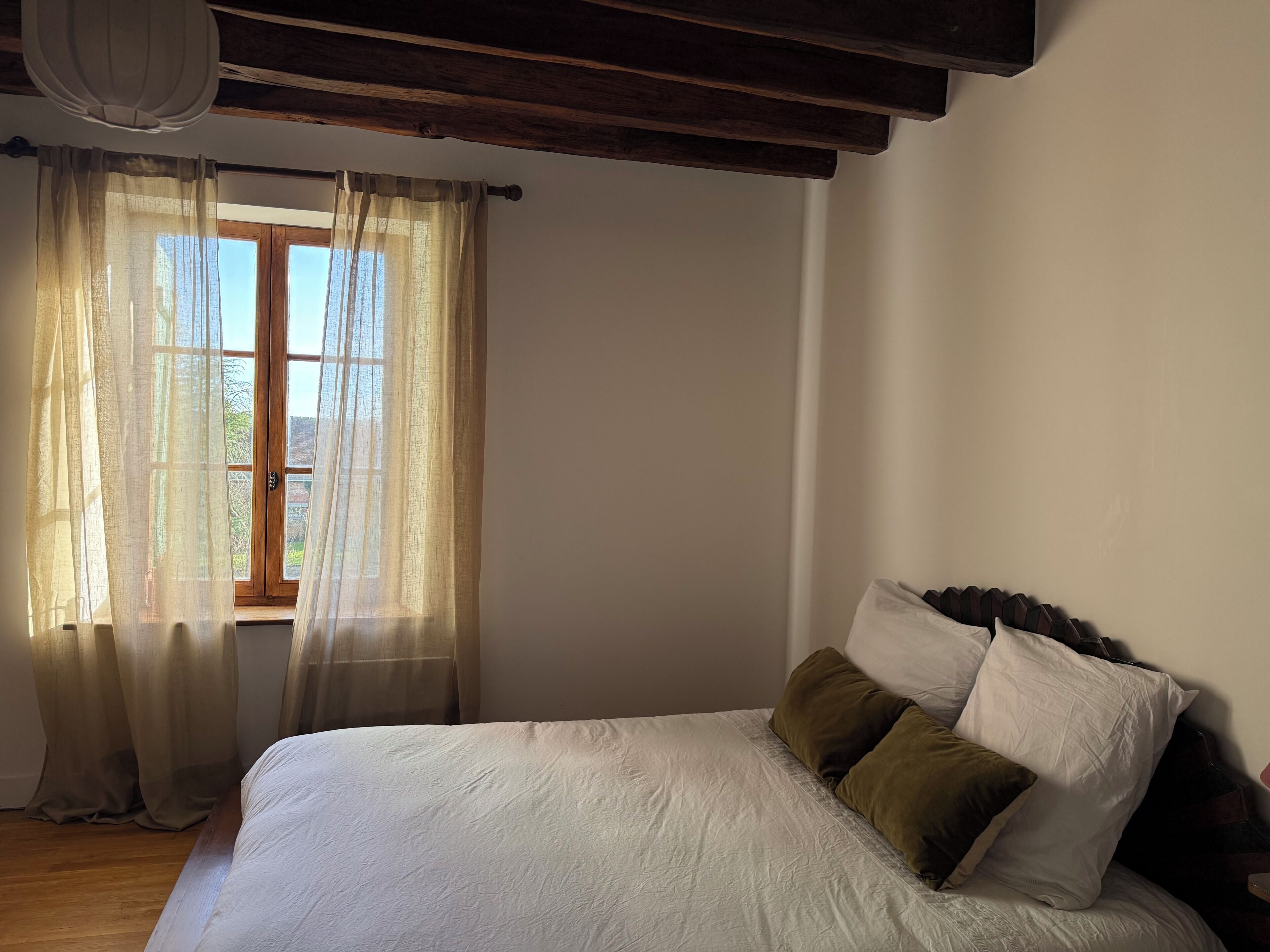 Une chambre avec un plafond en bois, un lit avec une literie blanche et marron, une fenêtre avec des rideaux légers, et une vue sur la verdure à l’extérieur.