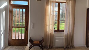 Una habitación con puerta de madera, ventana con cortinas translúcidas, un radiador y un pequeño banco de madera.