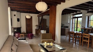 Una sala de estar con sofá, mesa de centro de vidrio, vigas de madera, un área de comedor con sillas y una cocina al fondo.