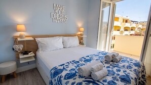 Une chambre d’hôtel avec un lit, deux lampes de chevet, un élément décoratif mural, une table de chevet avec un livre et un objet décoratif en forme de cœur, et une vue sur les bâtiments environnants.