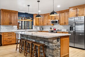 A kitchen with wooden cabinets, a large island with stools, a stainless steel refrigerator, and pendant lighting.