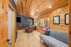 A log cabin interior with a wooden ceiling, a gray sofa, a loveseat, a leather chair, a wooden cabinet, and a flat-screen TV.