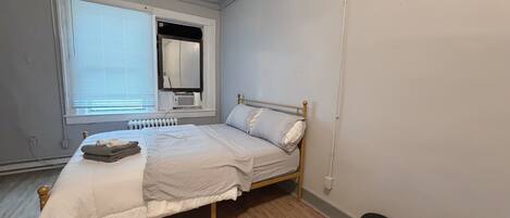 A bedroom with a bed, a radiator, a window with blinds, a mirror, and a trash bin.