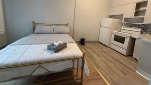 Una habitación pequeña y bien iluminada con una cama, kitchenette y un bote de basura.