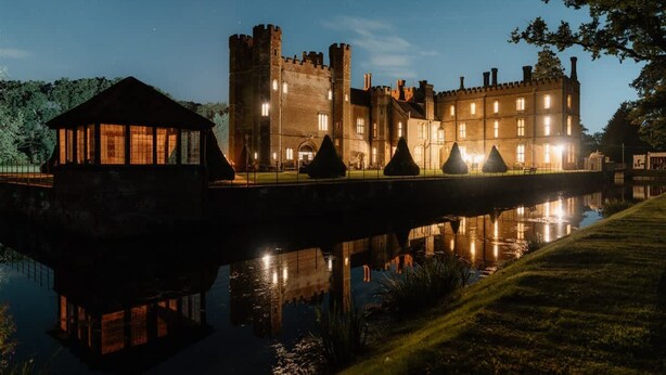 A castle-like building with a reflective water body in front, a small pavilion, and a bridge.