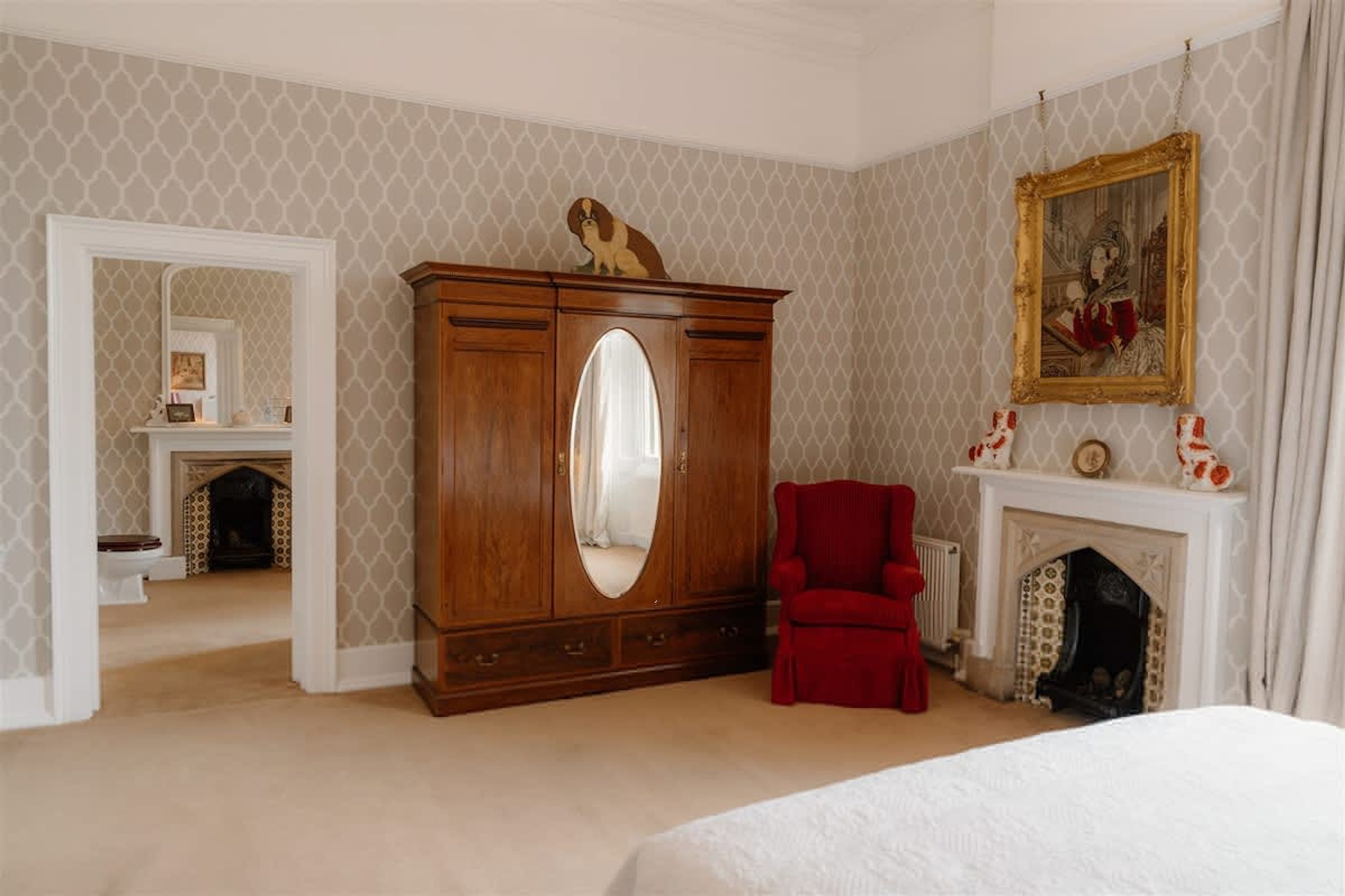 A bedroom with a large wooden wardrobe, a red armchair, a fireplace, and a large mirror.