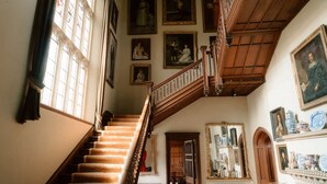 A grand staircase with ornate railings, a series of framed portraits on the walls, and a fireplace with decorative items.