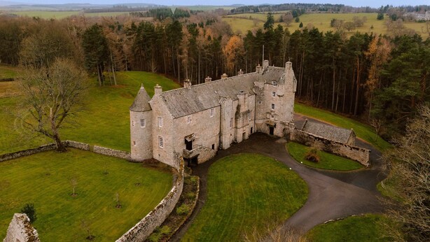 A large stone castle with multiple towers, surrounded by a well-maintained lawn and a forested area. - Lovingly restored castle in the Scottish Borders (Oxnam)