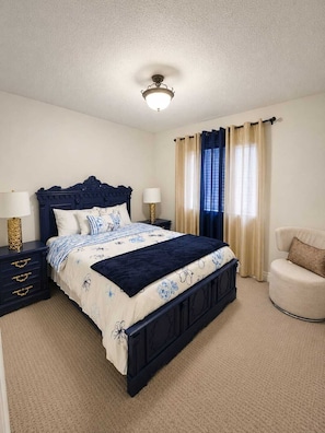 Un dormitorio con cama azul oscuro, ropa de cama blanca y azul estampada, una mesita de noche con lámpara, una silla y una ventana con cortinas.