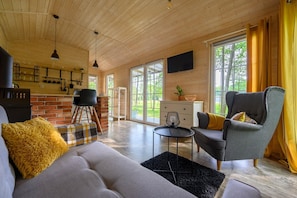 A cozy living room with a sofa, armchair, and a small table. There is a fireplace, a television mounted on the wall, and large windows with curtains. - Comfortable Holiday Home no. 2B With a Fireplace (Darlowo)
