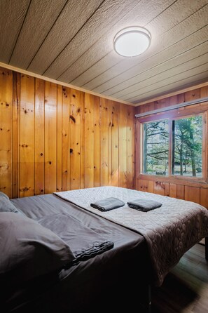 A wooden-paneled room with a bed, a window, and a ceiling light. - 3-Bedroom Cottage #9 (Nobel)