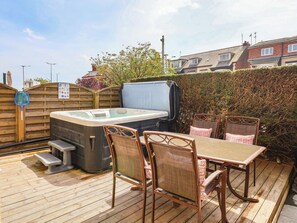 Ein Whirlpool mit Tisch und Stühlen auf einer Holzterrasse, umgeben von einem Holzzaun und einem Heckenstreifen.