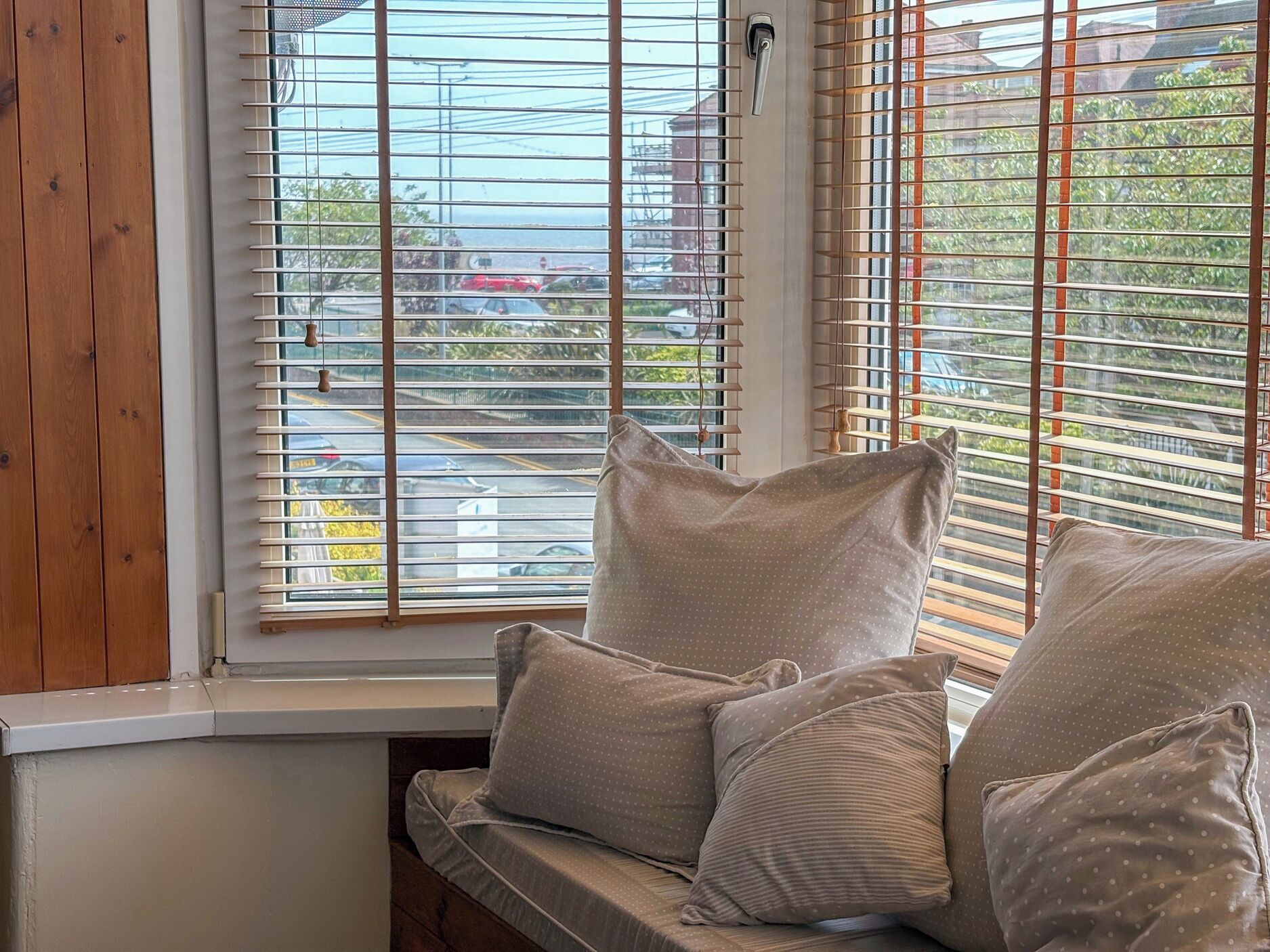 A window with horizontal blinds, a wooden bench with cushions, and a view of a street and buildings.