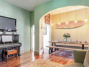 Ein Wohnzimmer mit einem Klavier, einem Esstisch mit Bänken, einem großen Gemälde und einer Leuchte.