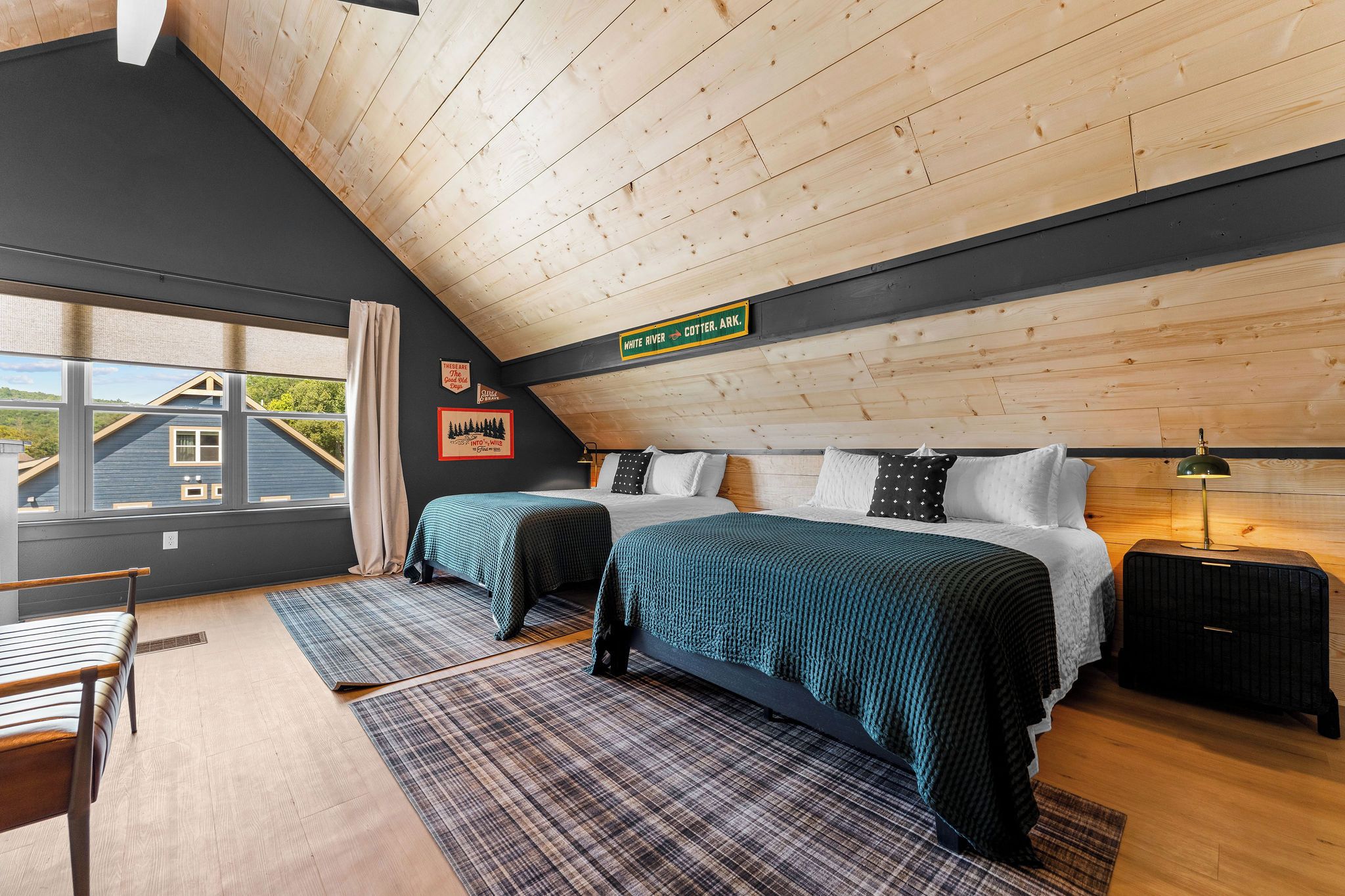 Un dormitorio con techo inclinado, una cama con manta verde, una mesita de noche, una lámpara y vista a una casa a través de una ventana.