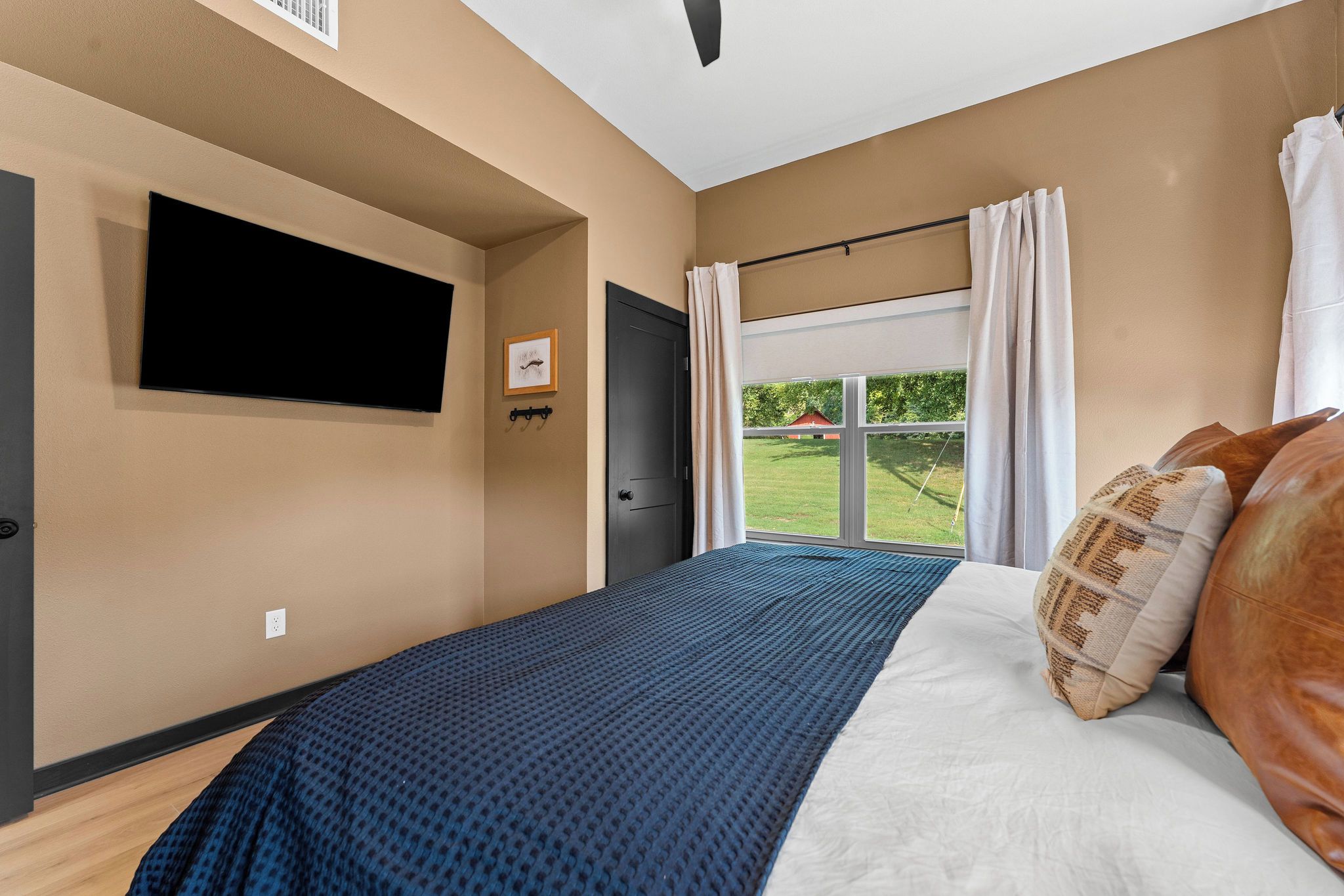 A bedroom with a bed, a flat-screen TV mounted on the wall, a door, and a window with curtains.