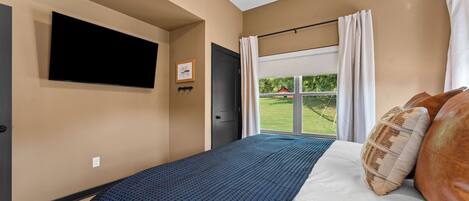 A bedroom with a bed, a flat-screen TV mounted on the wall, a door, and a window with curtains.